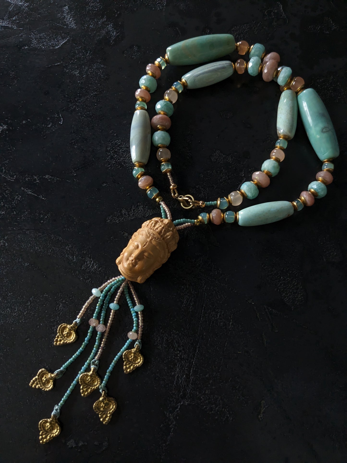 Le collier Bodhisattva de MUDRA est fait à la main avec de l'agate terra aqua, de l'amazonite, de la pierre de lune pêche et de la calcédoine. Il est orné d'une sculpture centrale de bodhisattva délicatement taillée dans du buis.