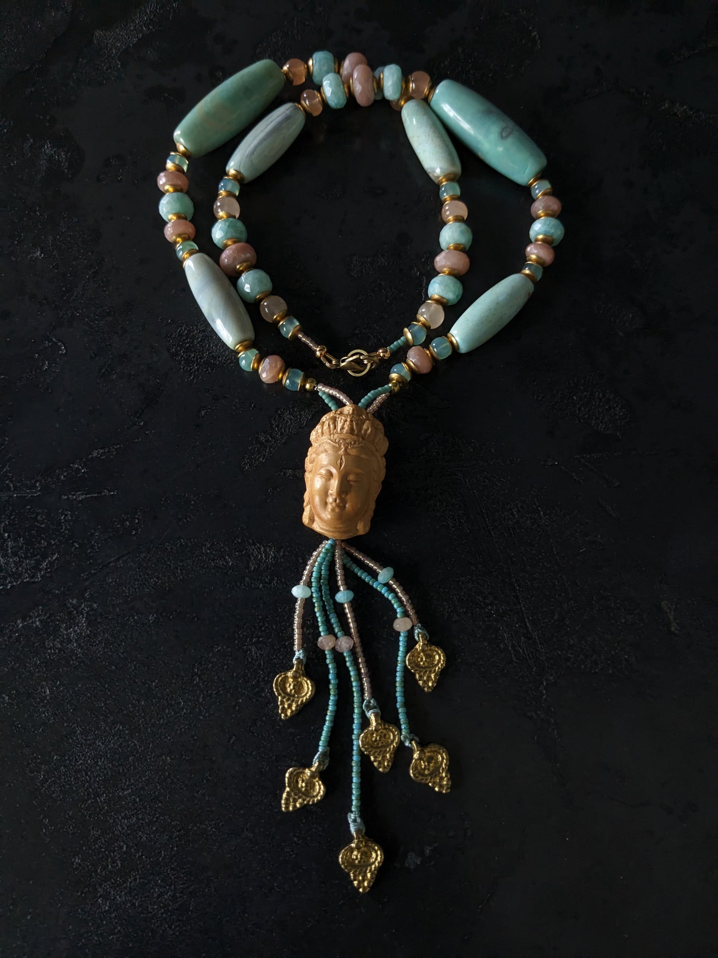 Le collier Bodhisattva de MUDRA est fait à la main avec de l'agate terra aqua, de l'amazonite, de la pierre de lune pêche et de la calcédoine. Il est orné d'une sculpture centrale de bodhisattva délicatement taillée dans du buis.