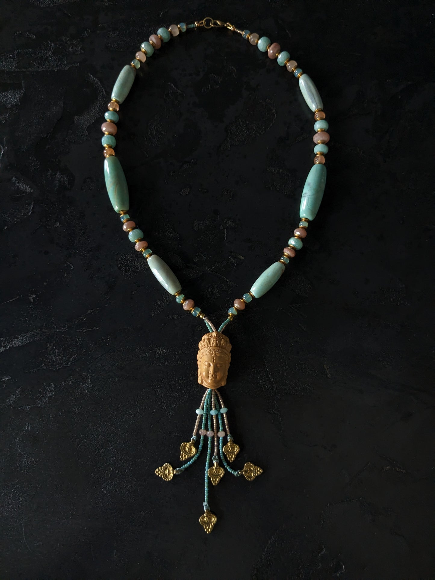 Le collier Bodhisattva de MUDRA est fait à la main avec de l'agate terra aqua, de l'amazonite, de la pierre de lune pêche et de la calcédoine. Il est orné d'une sculpture centrale de bodhisattva délicatement taillée dans du buis.