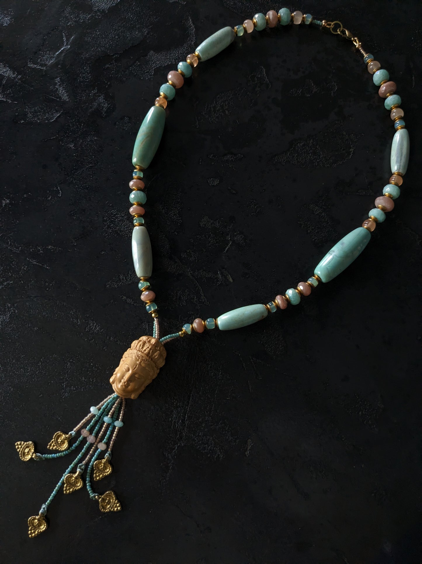 Le collier Bodhisattva de MUDRA est fait à la main avec de l'agate terra aqua, de l'amazonite, de la pierre de lune pêche et de la calcédoine. Il est orné d'une sculpture centrale de bodhisattva délicatement taillée dans du buis.