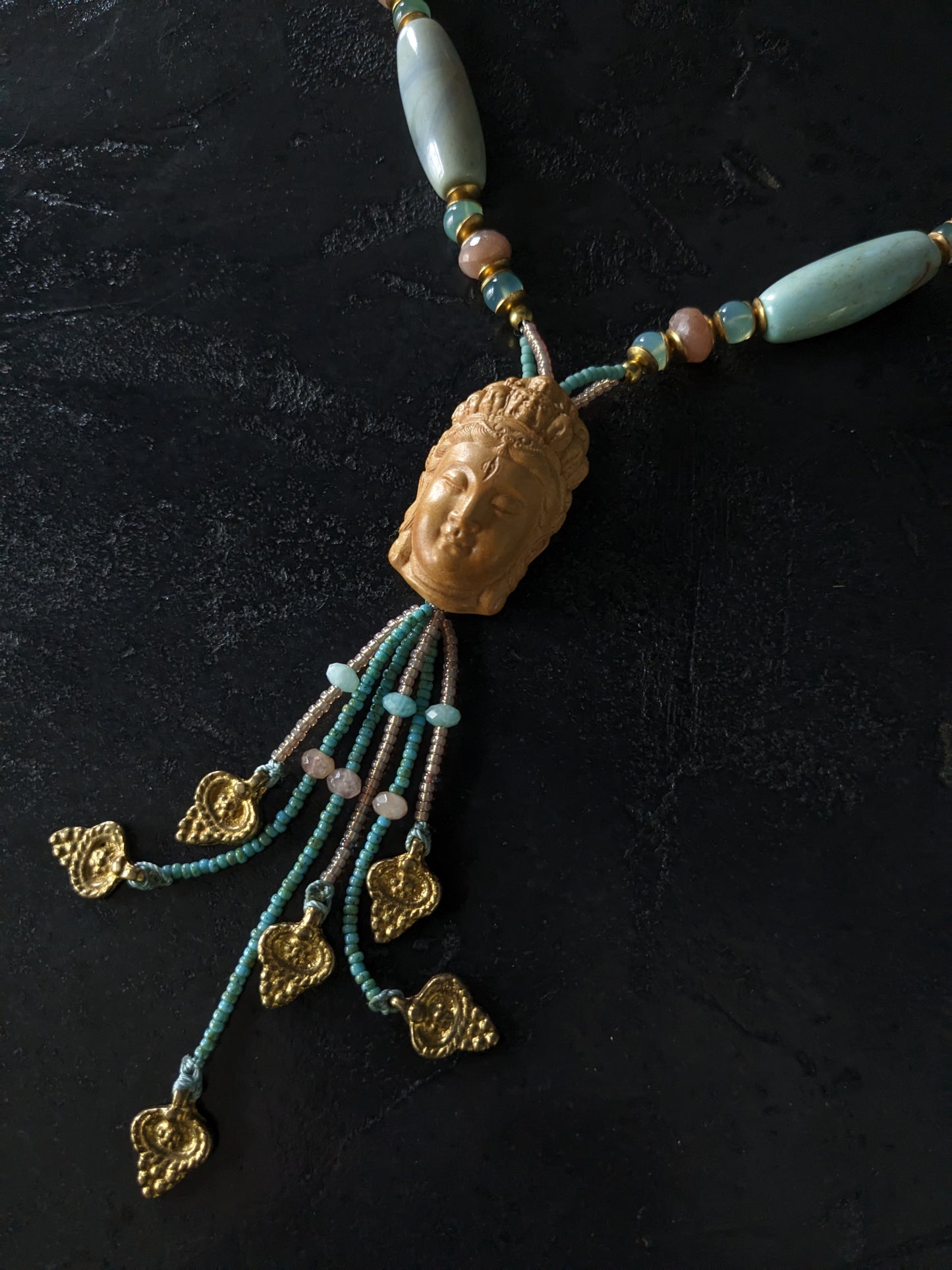 Le collier Bodhisattva de MUDRA est fait à la main avec de l'agate terra aqua, de l'amazonite, de la pierre de lune pêche et de la calcédoine. Il est orné d'une sculpture centrale de bodhisattva délicatement taillée dans du buis.