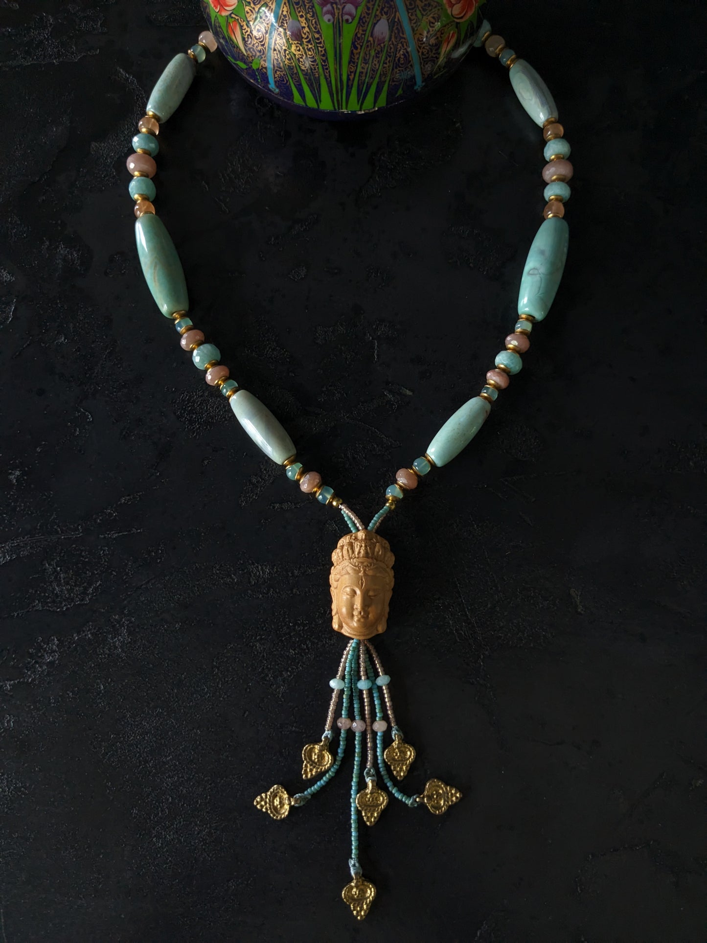Le collier Bodhisattva de MUDRA est fait à la main avec de l'agate terra aqua, de l'amazonite, de la pierre de lune pêche et de la calcédoine. Il est orné d'une sculpture centrale de bodhisattva délicatement taillée dans du buis.