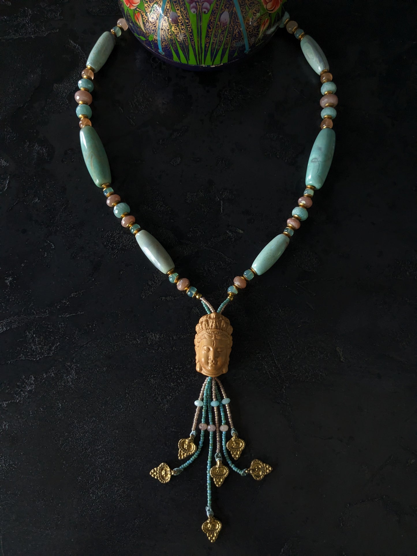 Le collier Bodhisattva de MUDRA est fait à la main avec de l'agate terra aqua, de l'amazonite, de la pierre de lune pêche et de la calcédoine. Il est orné d'une sculpture centrale de bodhisattva délicatement taillée dans du buis. 