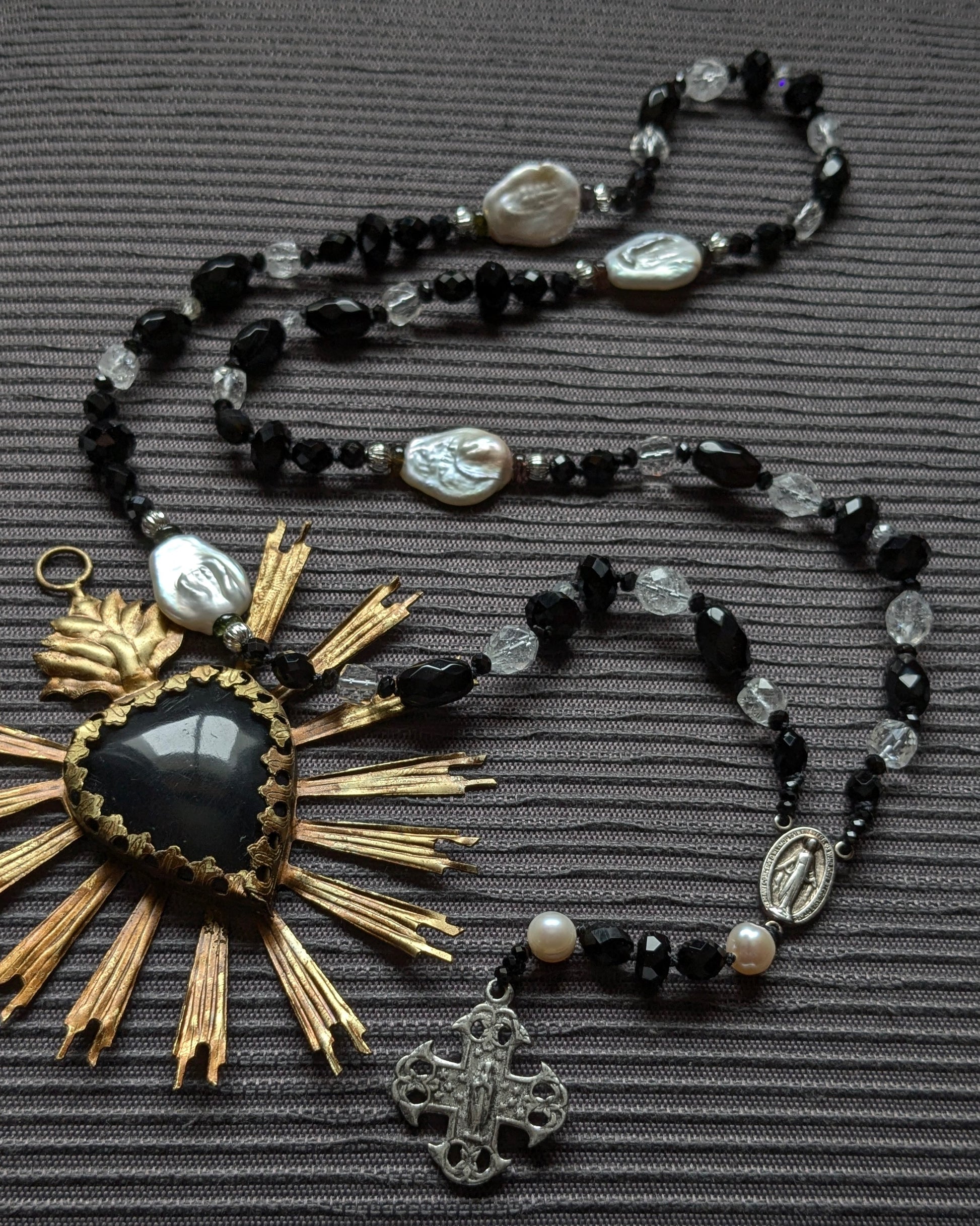 Chapelet artisanal en onyx noir facetté, obsidienne, tourmaline noire et cristal de roche, ponctué de perles d'eau douce baroques blanches irrégulières. Crucifix argenté ajouré et médaille de la Vierge Marie, posé sur un tissu gris texturé avec un soleil en laiton doré vintage en arrière-plan.
