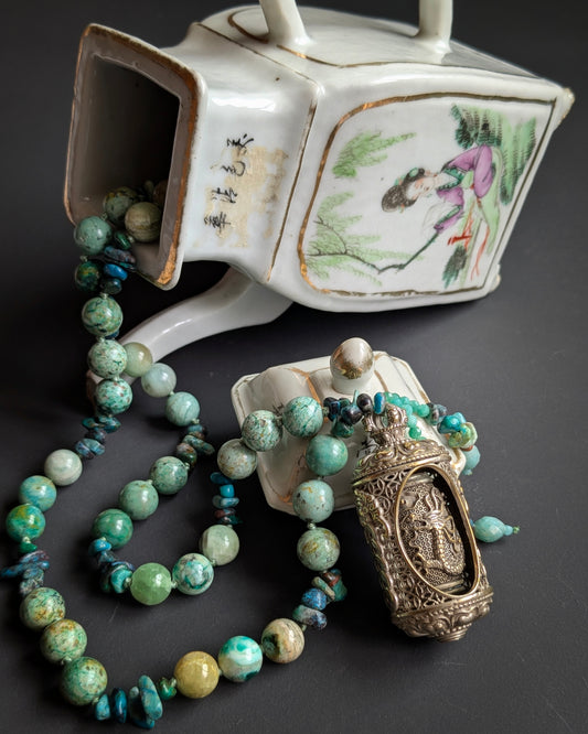 Collier de pierres naturelles vertes et bleues et pendentif moulin à prières ciselé, posés près d'une théière ancienne en porcelaine