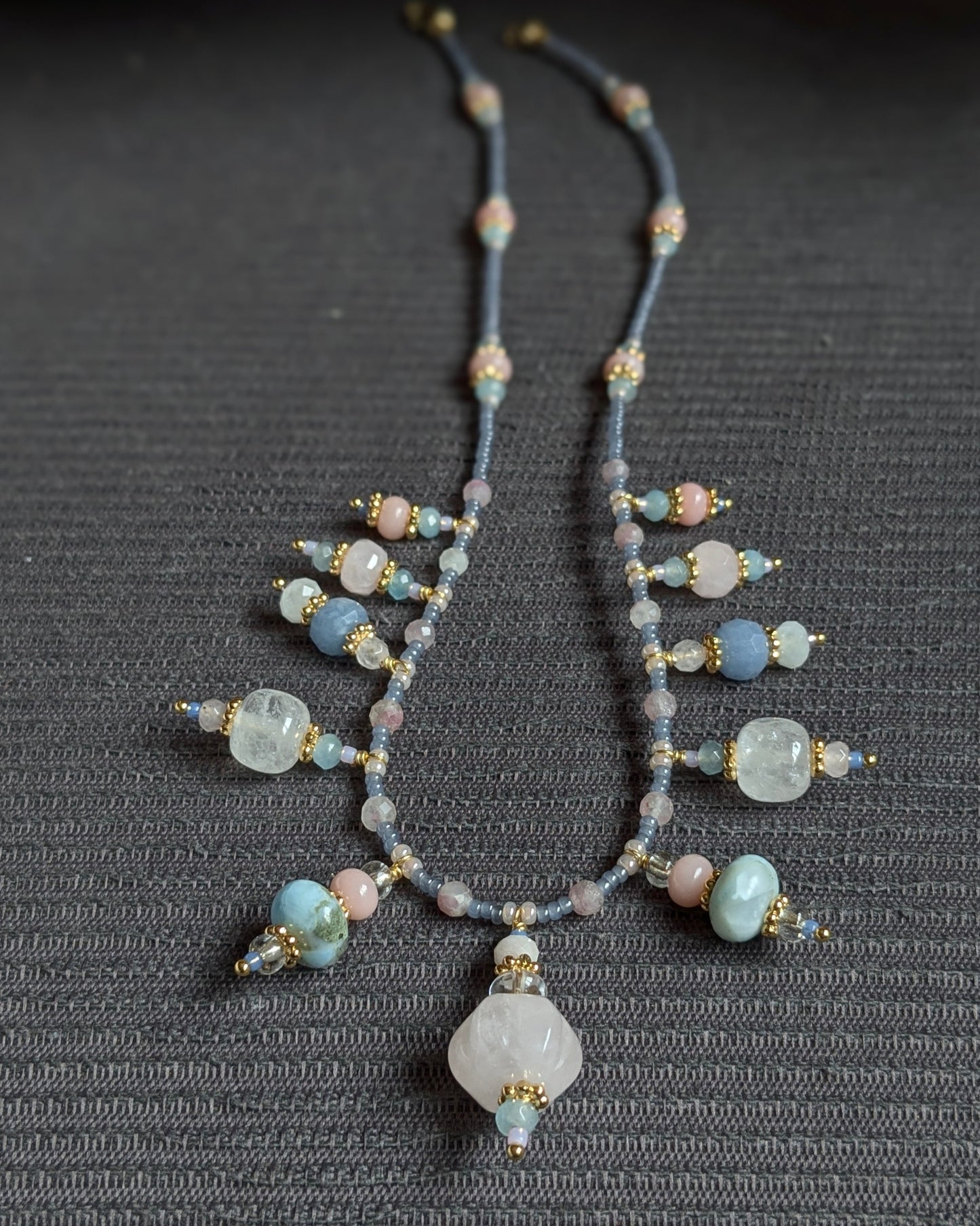 Le collier Prishaara exposé à plat, révélant le rythme de ses pierres entre quartz rose sculpté, éclats de larimar et transparence du cristal de roche.
