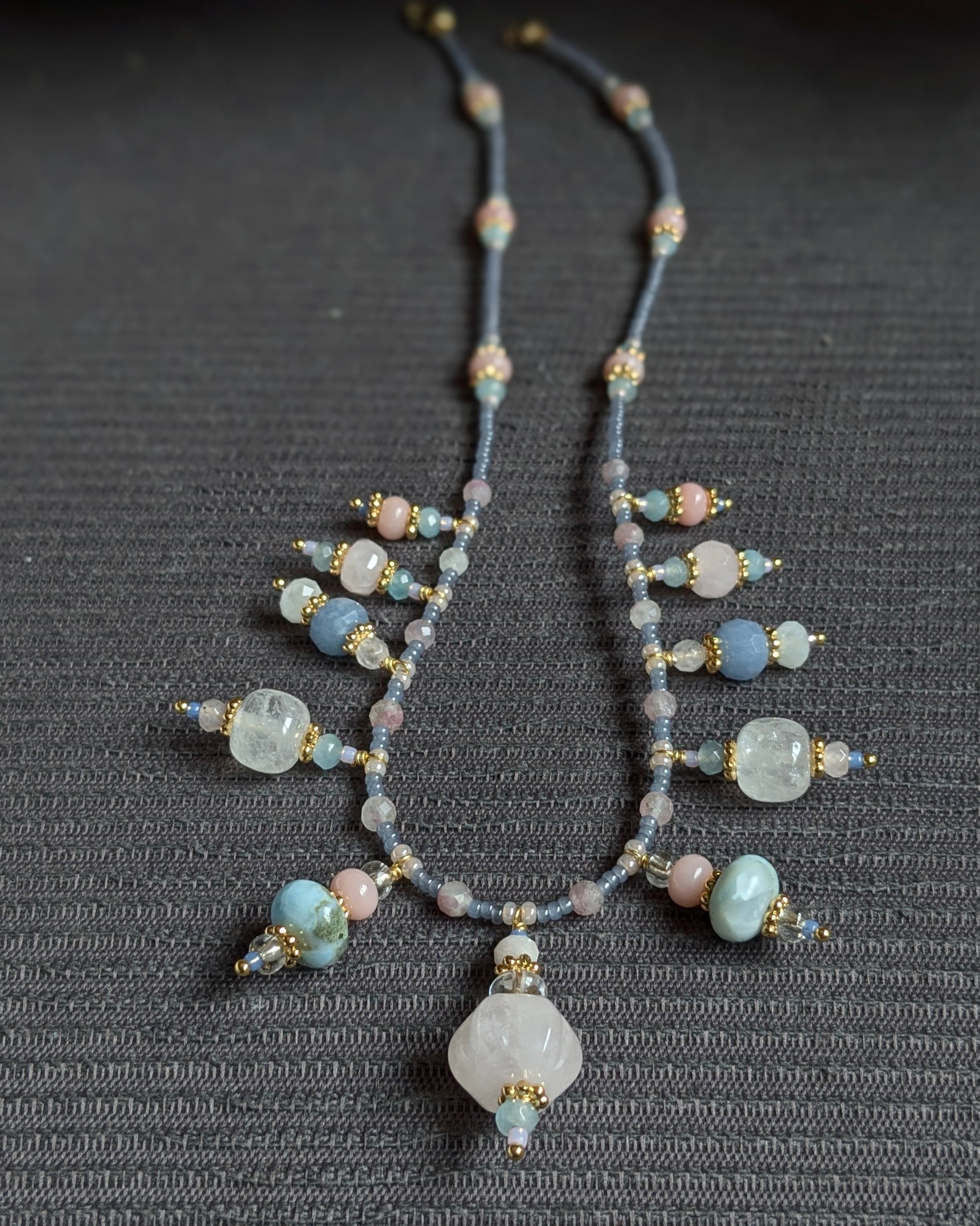 Le collier Prishaara exposé à plat, révélant le rythme de ses pierres entre quartz rose sculpté, éclats de larimar et transparence du cristal de roche.
