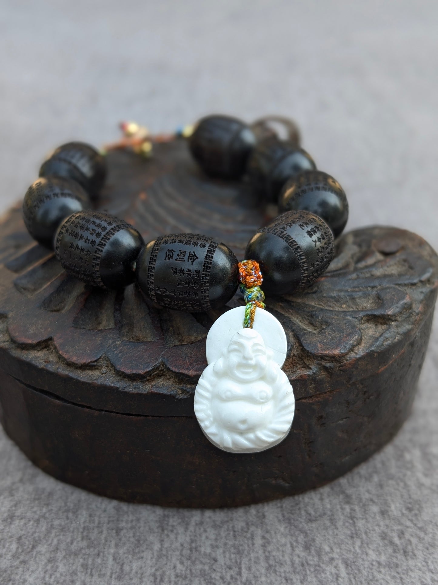 Détail du bracelet Norbu Mudra posé sur une boîte ancienne, gros plan sur les perles en santal noir gravées du Sûtra du Cœur et le pendentif Budai en coquillage blanc